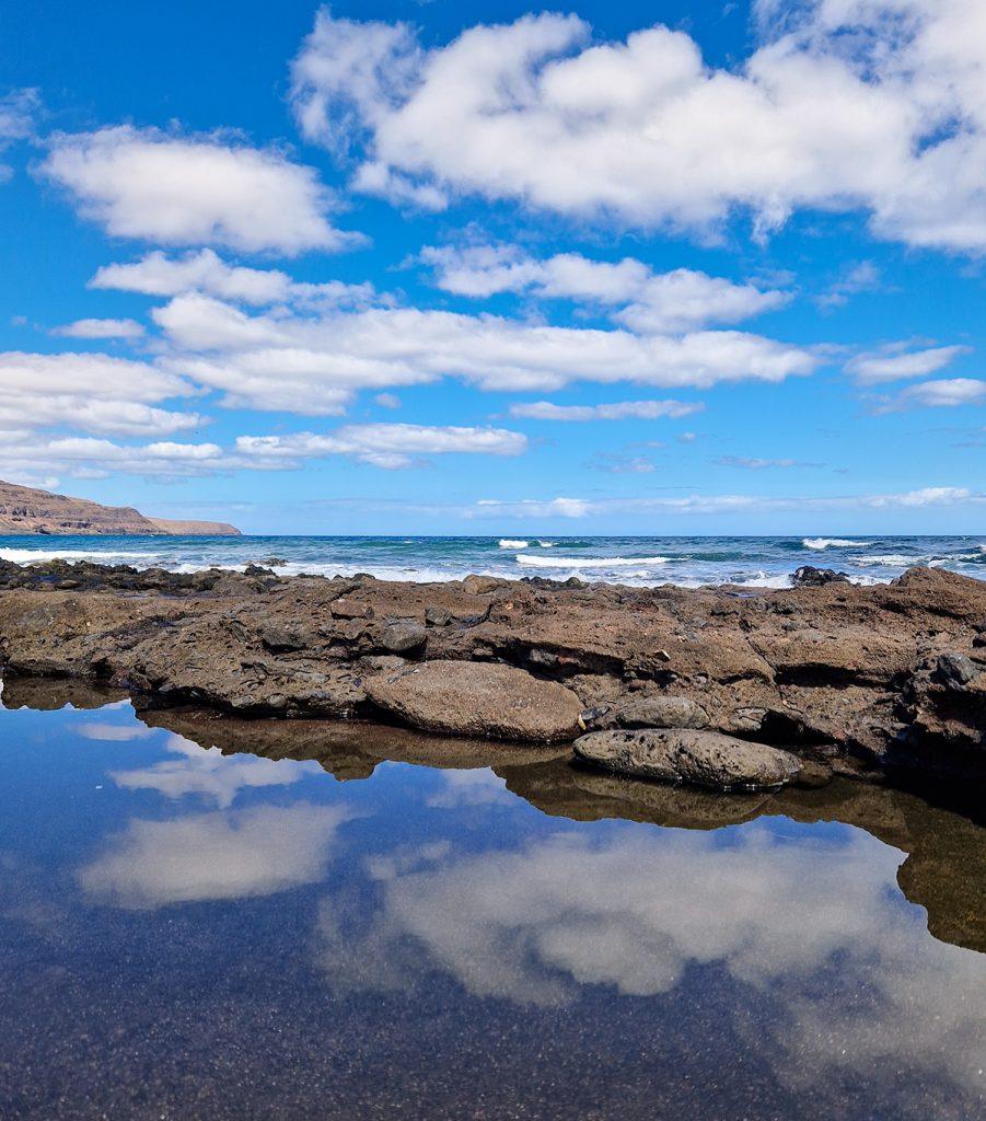 Charco natural de aguas tranquilas reflejando el cielo azul y nubes, con rocas volcánicas oscuras y el océano Atlántico al fondo, en la costa de Fuerteventura.